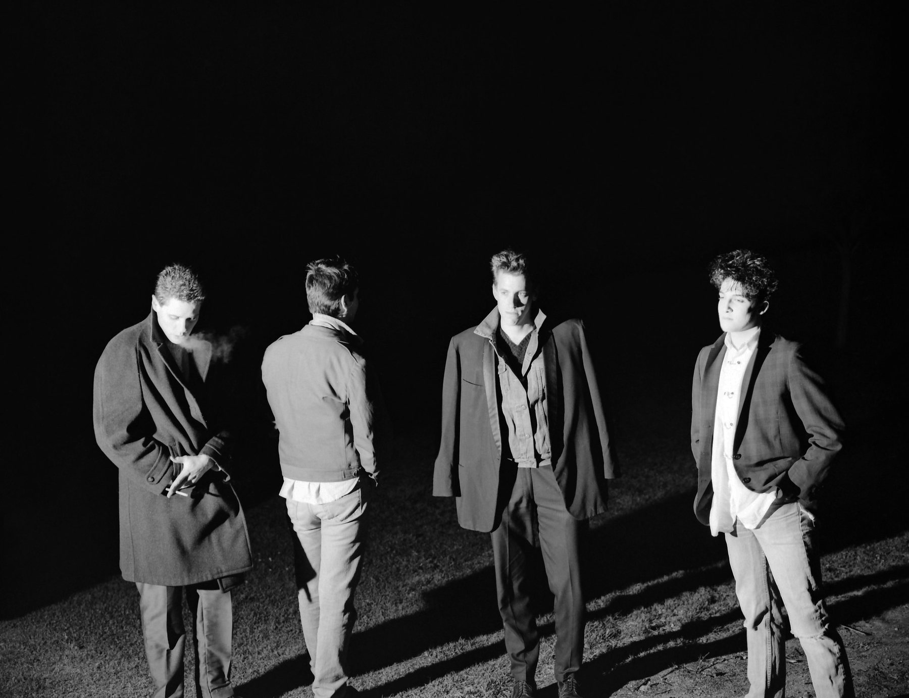 Kissing Jane: four young men in coats lit hard against a black night sky